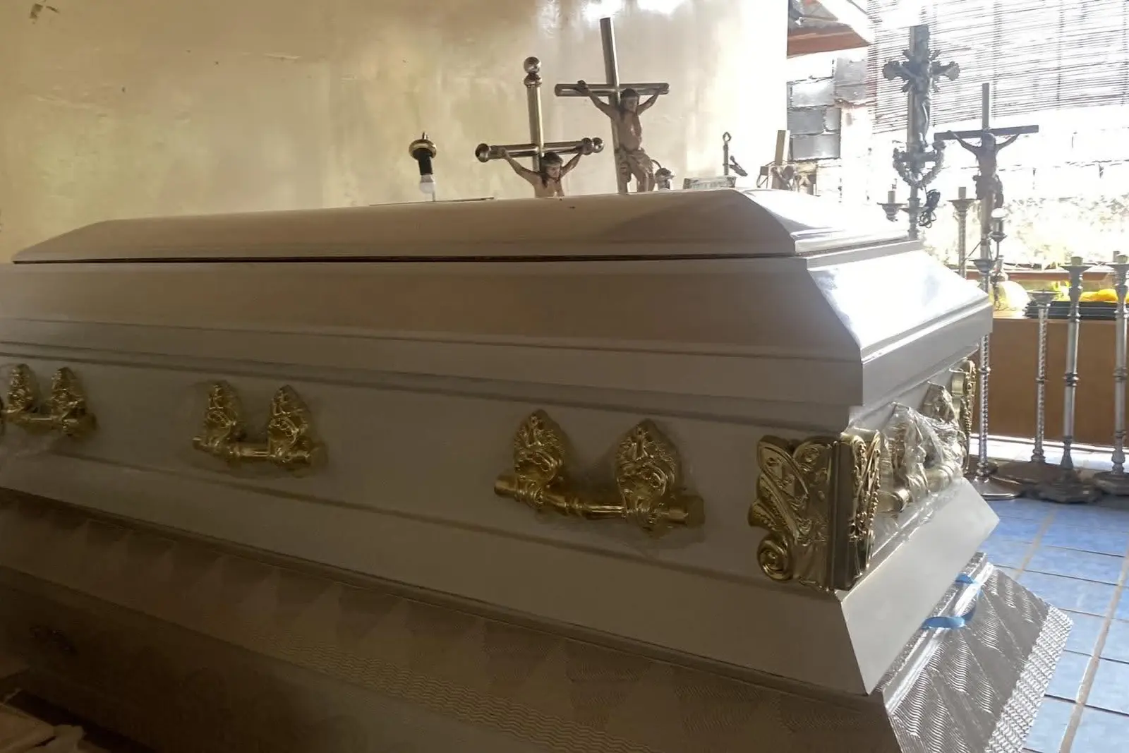 Illustrative sample coffin — A white casket with ornate gold handles and a closed full-glass lid, photographed in a chapel with crucifixes behind
