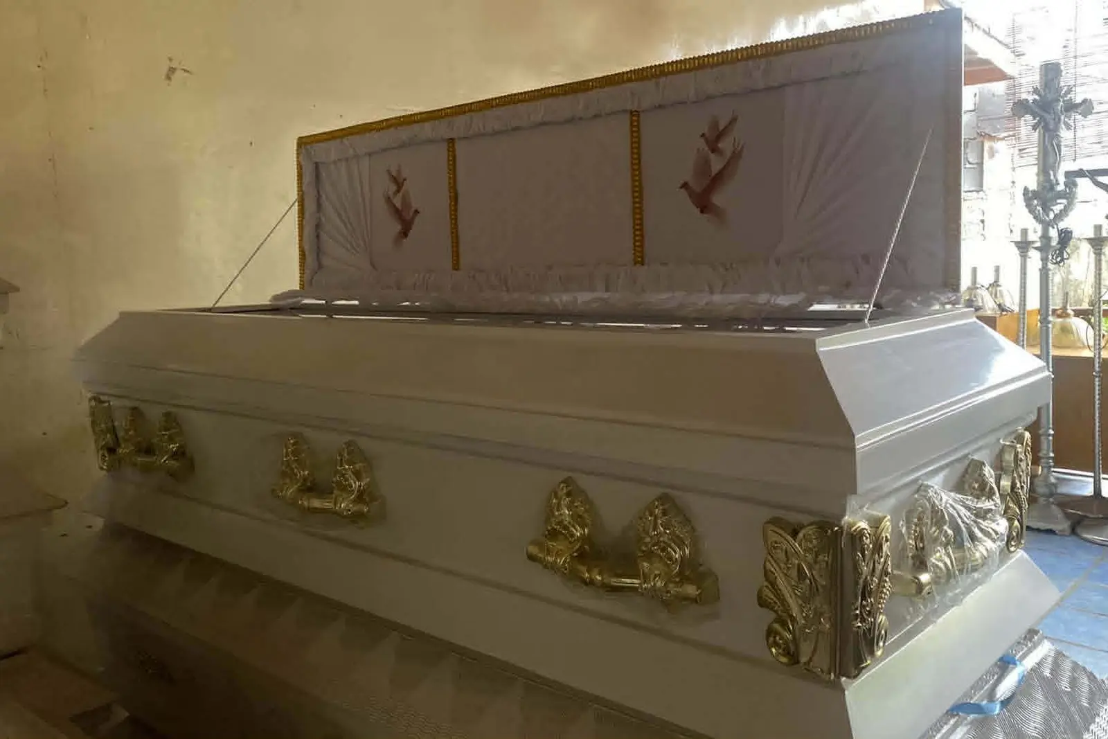 Illustrative sample coffin — A white casket with gold handles and a raised full-glass lid showing a white quilted interior with dove embroidery, in a chapel