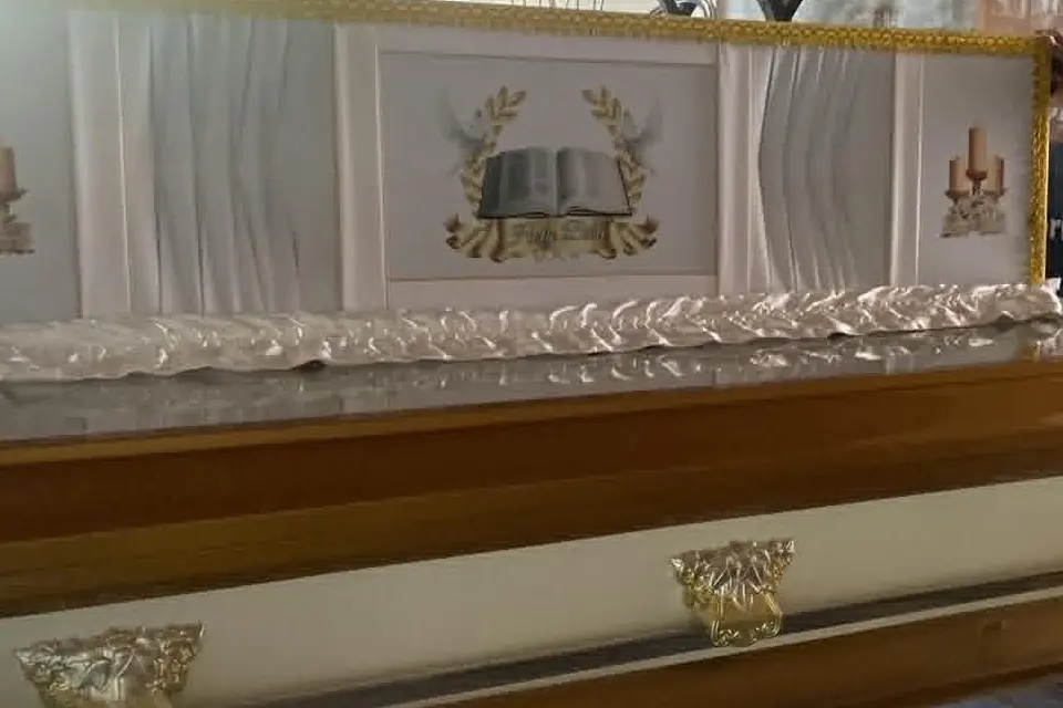 Illustrative sample coffin — The raised full-glass lid of a wood-and-white casket, its white interior embroidered with an open bible between candelabra, above a gold side handle