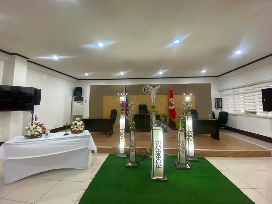 A chapel hall with a white-draped table, tall candle pedestals on a green carpet runner and a panelled platform behind
