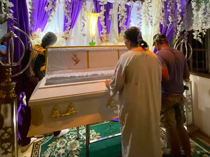 A white casket with gold handles in a purple-draped alcove under white flower garlands, with two people arranging the set-up