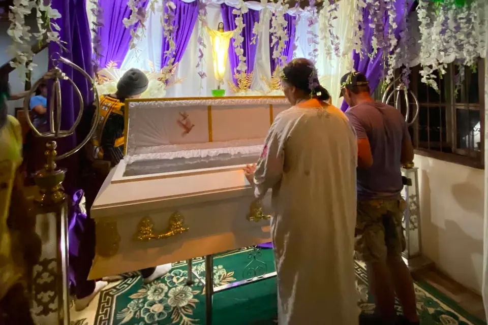 A white casket with gold handles in a purple-draped viewing room the office prepared, under garlands of white flowers