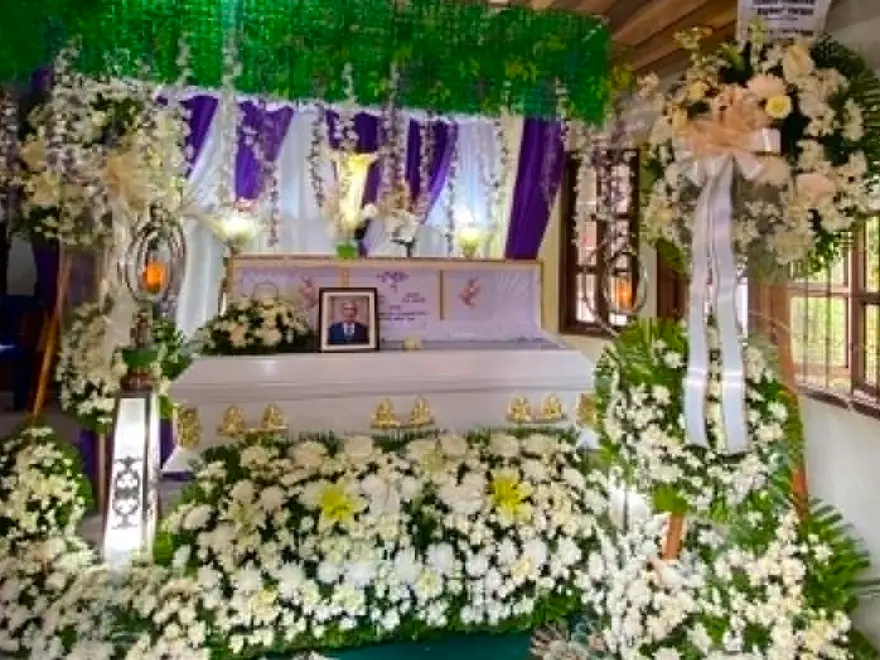 A finished wake set-up — a white casket banked in white flowers under green foliage and purple drapes, with a framed portrait